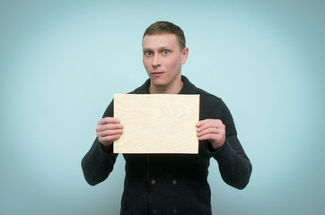 Surprised man is holding in front of himself a wooden board with copy space isolated on blue background.