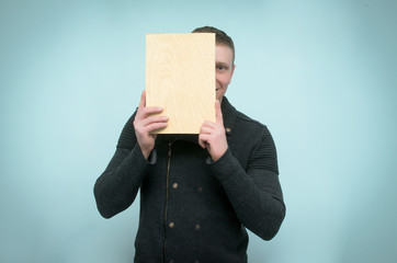 Happy man hides his face behind a wooden plaque plate in his hands and peeks out from there isolated on blue background.