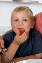 close up portrait of young 3 years old boy with his mother at home kitchen eating fruit or tomato enjoying happy in healthy nutrition