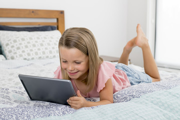 Obraz na płótnie Canvas sweet and beautiful blond 6 or 7 years old young girl lying on bed smiling happy using the internet on digital tablet pad watching and having fun