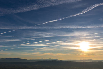 Sunset over distant mountains, with blue sky full of white clouds and vapor trails