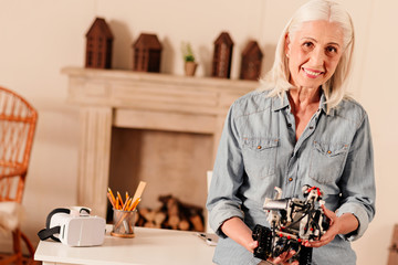 Contemporary retirement. Waist up shot of retired woman looking into the camera with a cheerful mile on her face while standing at her table and holding a robotic machine.