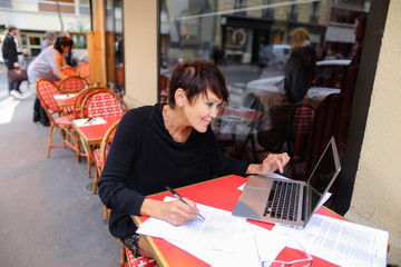 middle aged female student using laptop for writing a research work. Woman sitting at red table in...
