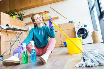 young woman in rubber gloves holding mop and sitting on floor while cleaning house