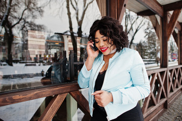 Stylish african american plus size model with mobile phone at hand against wooden cafe at winter...