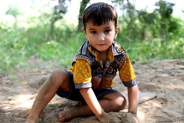 Cheerful child of Indian ethnicity sitting in soil & playing with sand outdoor in the nature.