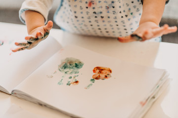 toddler girl in drawing apron draws fingers at home, close up of colored hands