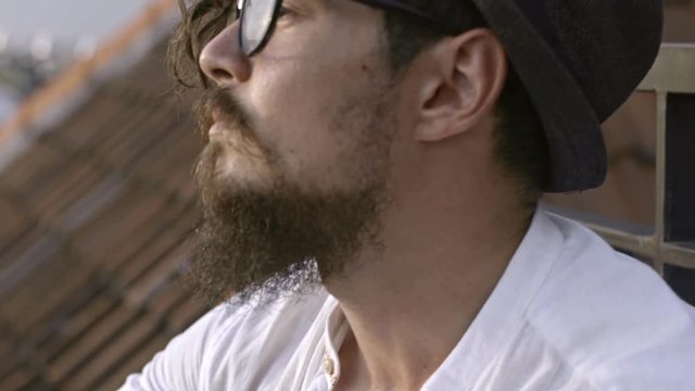 Thoughtful bearded man in eyeglasses and hat enjoying view from rooftop and then looking at camera
