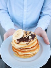 Man is holding plate with freshly made homemade pancakes with chocolate nougat, grounded peanuts and walnut, sliced banana. Sweet breakfast, food for freakfast. Gluten free pancakes. Closeup