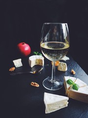 Delicious assorted cheese with fresh fruits on a black stone background. White wine glasses, Gorgonzola, blue Roquefort, Brie cheese with honey. Appetizers, sliced cheese on a plate.