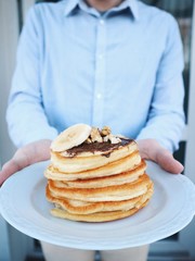 Freshly made delicious pancakes with chocolate nougat, bananas, nuts on a white plate. Man holding a plate with pancakes. Hands holding plate. Tasty homemade breakfast. Healthy breakfast ideas.