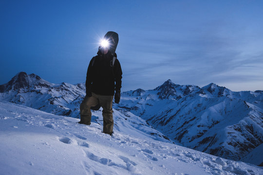 Man with headlamp and backpack wearing ski wear standing in front of amazing winter mountain view. Traveler climb at night on the snowy mountain. Ski tour