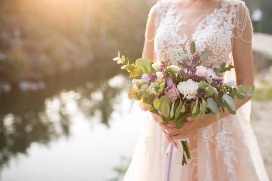 The bride in a beige wedding dress holding a lush bridal bouquet of lilac roses and a lot of greenery. Stylish wedding bouquet on the sunset background