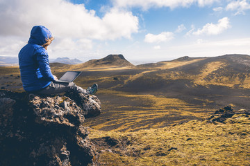 Young woman hiking a mountain in Iceland and sitting on the rock while using a laptop at sunset...