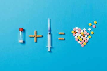 Pills bottle plus empty syringe needle equals medication colorful round tablets in form of heart isolated on blue background. Concept of treatment, choice healthy lifestyle. Copy space advertisement.