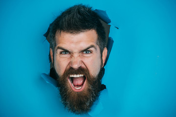 View of male face through hole in blue paper. Angry bearded man making hole in paper. Bearded man...
