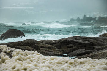 Crashing waves onto rocks on a misty day with Camps Bay, Cape Town in the background.