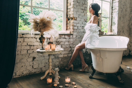 A beautiful bride in her underwear and a large men's white shirt in shoes sits on the bathroom by the window, next to a small white table with candles in glasses and lush feathers in metal vases