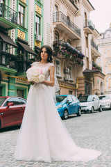Obraz premium The bride in a wedding dress is standing on the road against the background of a street with a bouquet in her hands and raises her hand