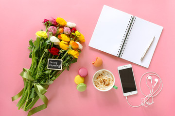 Festive morning concept buttercup flowers with note good morning, cup of cappuccino, cake makaron, mobile with headphones, notebook, pen on the pink background.