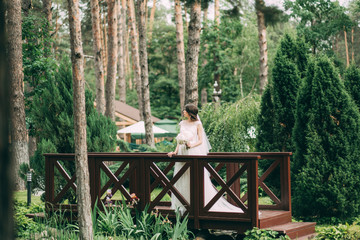 Fototapeta na wymiar Bride in wedding dress and holding a bouquet in hands is standing on a wooden brown bridge in nature.