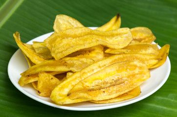 Fried banana chips on white plate with banana leaf background.