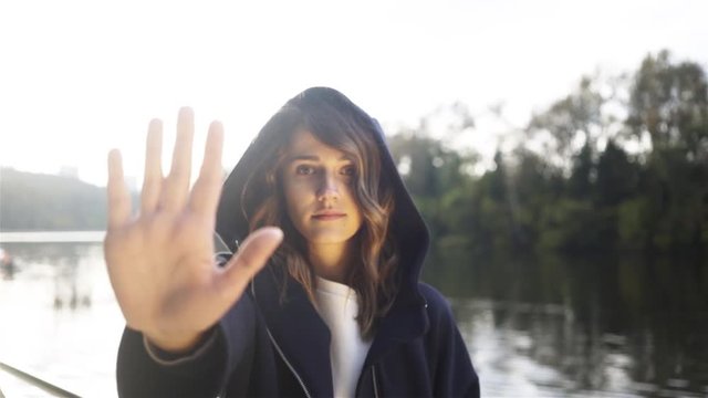 Pretty young brunette in a black hoodie showing a no sign standing on a river bank on a sunny autumn day. Zoom out slow motion medium shot