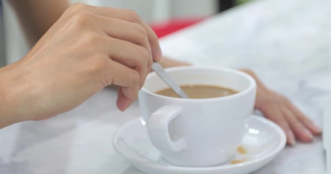A cup of coffee, young asian white woman by hands is mixing coffee with metallic spoon in white ceramic cup on white marble table.