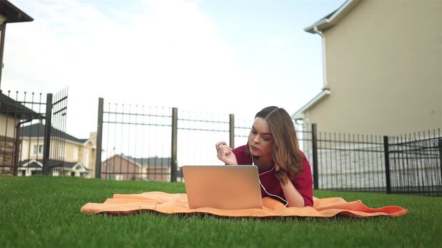 Young woman in a red sweatshirt lying in her backyard with a laptop and headphones on a summer day. Left to rigth pan real time medium shot