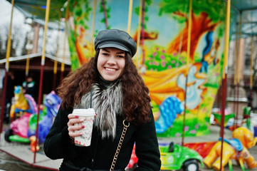 Fototapeta na wymiar Curly mexican girl in leather cap and plastic cup of coffee at hand against carousel.