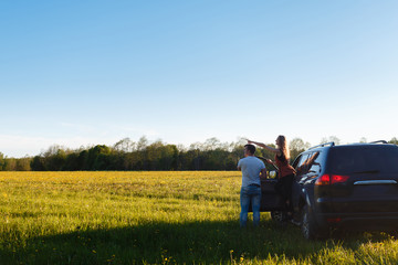 A loving couple watch the sunset, standing leaning against an black car. The young man shows to his girlfriend sunset standing near SUV. Place for text.