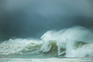 Huge waves crashing on a beach on a misty dark day with mountain in the background.