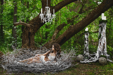 Fairy tale picture - beautiful sexy brown-haired model girl wears lingerie, rests in a huge nest in the green forest