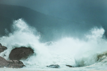 Huge waves crashing on rocks on a misty dark day with mountain in the background.