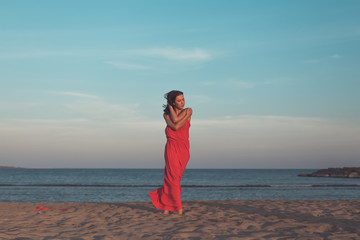 attractive young woman in red dress is standing  on the beach at suset