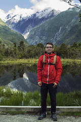 Fototapeta na wymiar Hombre joven posando frente a paisaje de picos de montañas nevados y verdes con cielo nublado reflejado en un lago en Nueva Zelanda
