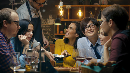 In the Bar Waiter Holds Credit Card Payment Machine and Beautiful Woman Pays for Her Order with Contactless Mobile Phone Payments System. She's Surrounded by Dear Friends and Has Time of Her Life.