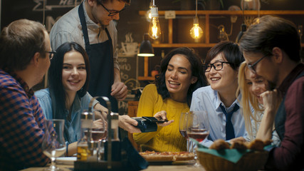 In the Bar Waiter Holds Credit Card Payment Machine and Beautiful Woman Pays for Her Order with Contactless Mobile Phone Payments System. She Has Good Time with Her Friends.