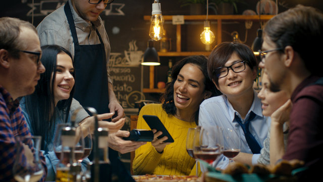 In the Bar Waiter Holds Credit Card Payment Machine and Beautiful Woman Pays for Her Order with Contactless Mobile Phone Payments System. She Has Good Time with Her Friends.