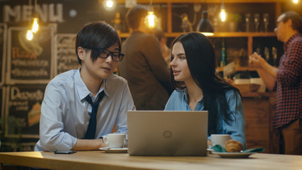 Handsome Asian Man and Beautiful Caucasian Young Woman Sitting in the Cafe Work on a Laptop Computer. In the Background Other Customers in the Stylish Environment.
