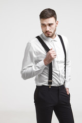 Fashionable hipster guy with stubble and stylish haircut posing at blank studio wall, looking away with mysterious expression. Handsome young bearded male dressed in formal wear pulling suspenders