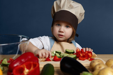 Children, food, nutrition and healthy lifestyle concept. Adorable joyful 5 year old baby girl in chef uniform cutting various vegetables on cooking board while making vegetarian lasagna or soup