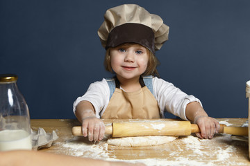 Happy cute little girl flattening pastry using rolling pin while helping mother cooking pie for...