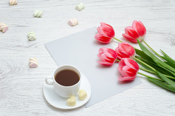 Fototapeta na wymiar Pink tulips, mug of coffee and sheet of paper. Light wooden background.
