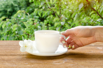 Female hand with white mug of tea and jasmine on a wooden table. Close up