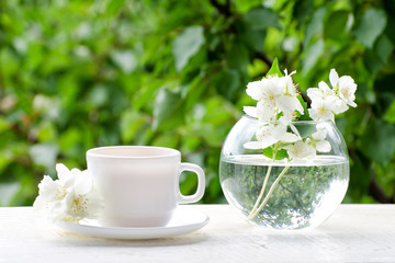 White mug of tea and a vase with jasmine on a wooden table, greens on the background