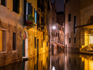 Fototapeta na wymiar Calm water flows along a canal under a bridge in Venice amidst old buildings illuminated at night