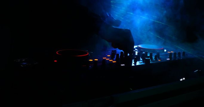 DJ Spinning, Mixing, and Scratching in a Night Club, Hands of dj tweak various track controls on dj's deck, strobe lights and fog, selective focus, close up. Slider Shoot