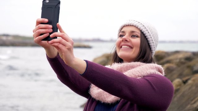 Happy beautiful woman in winter in front of the ocean taking selfie picture with cell phone