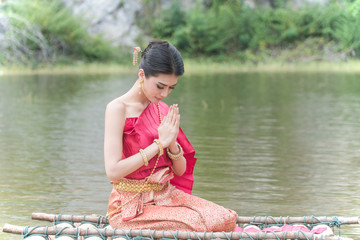 Portrait young woman wearing traditional thai red and purple and gold jewelry in the water park.Beautiful female sitting with hands clasped (Sawasdee -Thai greeting) on a raft floating in the lake.